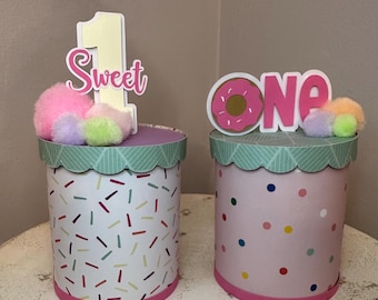 May include: Two pink and white donut-themed party favor boxes with a pink and white sprinkle pattern and a pink and white polka dot pattern. The boxes are topped with a pink and white donut and the words "one" and a pink and white number one with the words "sweet".