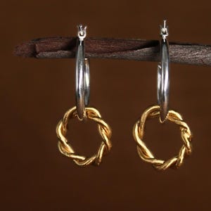 May include: A pair of hoop earrings. The top portion of the hoops is silver, while the bottom features a gold-colored, woven design. The earrings hang against a brown background.