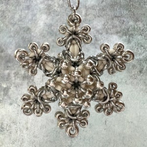 May include: A silver-colored snowflake pendant with a delicate design. The pendant features a central cluster of off-white beads surrounded by intricate silver wirework, forming six symmetrical points. The pendant hangs from a thin silver chain.