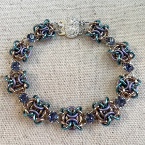 May include: A handcrafted bracelet with chainmaille links in teal, lavender, and bronze. The bracelet has small, round, purple gemstones and a silver clasp. The chainmaille design forms a floral pattern. The bracelet is approximately 19 cm in length.