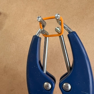 May include: Close-up of a blue and silver tool holding an orange square rubber band. The tool has two blue handles and silver metal components. The rubber band is centered within the tool's grip, showcasing its shape and color.