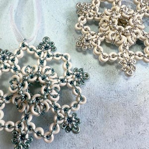 May include: Two snowflake ornaments. Each snowflake is white with silver-colored accents and a silver-colored chain. One snowflake has a white ribbon for hanging. The ornaments are on a light blue surface.