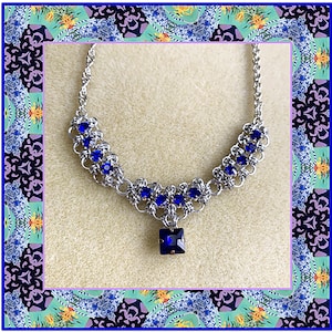 May include: A silver chain necklace with a blue crystal pendant and blue crystal accents. The necklace is on a beige background.