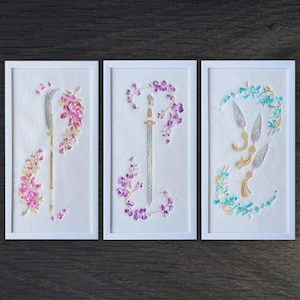 Peut inclure: Trois œuvres encadrées représentant des armes brodées et des motifs floraux. La première montre une hallebarde avec des fleurs roses, la seconde une épée avec des fleurs violettes, et la troisième, trois fers de lance avec des détails turquoise et dorés.