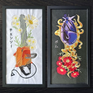 May include: Two framed embroidery pieces. The left piece on white fabric shows an orange chainsaw with white daisies and Japanese text. The right piece on black fabric features a purple, egg-shaped object with yellow flames and red flowers with Japanese text.