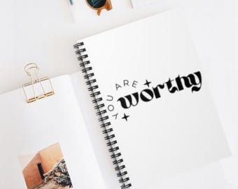 May include: White spiral-bound notebook with the words "ARE YOU WORTHY" in black script. The notebook is open, revealing a photograph and a gold binder clip. Sunglasses and a pen are also visible.