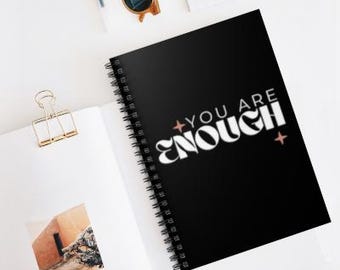 May include: A black spiral notebook with the text "You Are Enough" in white lettering. The text is stylized with a bold font and has a pink star on either side.