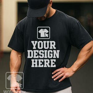 May include: Black t-shirt with the text "YOUR DESIGN HERE" in white block letters. The shirt features a graphic of a t-shirt and a cursor. The person in the image is wearing a black baseball cap.