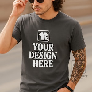 Può includere: T-shirt girocollo grigio scuro con la scritta bianca "YOUR DESIGN HERE". La maglietta presenta una grafica di una t-shirt e un cursore. Il modello indossa occhiali da sole e ha un tatuaggio sul braccio.