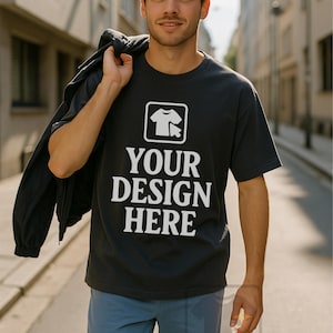 May include: A black t-shirt with the text "YOUR DESIGN HERE" in white. The shirt features a graphic of a t-shirt and a cursor. The person is wearing blue pants and holding a black jacket.