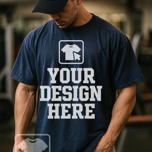 May include: Navy blue t-shirt with the text "YOUR DESIGN HERE" in white block letters. The shirt features a graphic of a t-shirt and a cursor. The person in the image is wearing a black baseball cap.