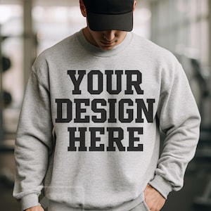 May include: A light gray sweatshirt with the text "YOUR DESIGN HERE" in bold black letters. The person in the image is wearing a black baseball cap and gray sweatpants. The sweatshirt has a crew neck and long sleeves.