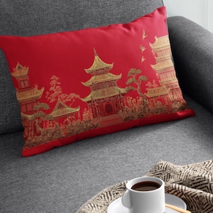 May include: A red decorative pillow with a gold Asian-inspired design featuring pagodas, trees, and figures. The pillow is rectangular and rests on a gray sofa. A cup of coffee sits on a saucer nearby.