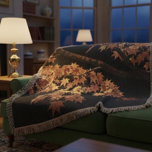 May include: A woven throw blanket draped over a green sofa. The blanket features a dark background with a design of autumn leaves in shades of orange and brown. The sofa is in a living room setting with a fireplace and lamps.