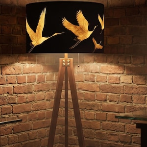 May include: A floor lamp with a tripod wooden base and a cylindrical black lampshade featuring a gold-colored graphic print of flying cranes. The lamp includes a metal pull chain switch and visible silver-toned hardware at the top of the legs.