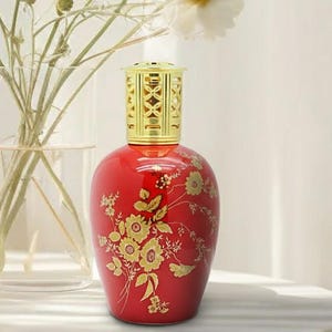 May include: A red decorative fragrance lamp with a gold-colored top and floral design. The lamp is on a white surface, with a vase of white flowers in the background.