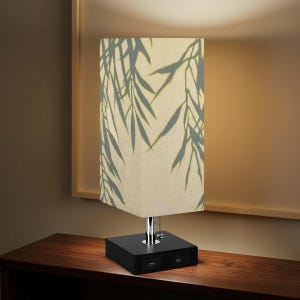 May include: A rectangular table lamp with a black base and a cream-colored shade. The shade features a gray bamboo leaf pattern. The base has two USB ports. The lamp is on a dark wooden surface.