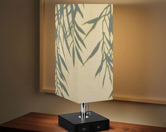 May include: A rectangular table lamp with a black base and a cream-colored shade. The shade features a gray bamboo leaf pattern. The base has two USB ports. The lamp is on a dark wooden surface.