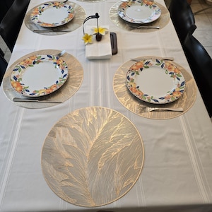 May include: A dining table set for a meal, featuring white plates with orange and floral designs, silver forks and knives, and gold-colored placemats. A white tablecloth and a napkin holder with a flower complete the setting.