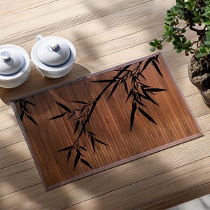 May include: A bamboo tea tray with a black bamboo leaf design, two white teacups with lids, and a small potted plant. The tray is rectangular and made of brown bamboo. The teacups are white with blue accents.
