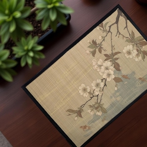 May include: Rectangular beige placemat with a black border, featuring a floral branch design with white blossoms and brown leaves. The placemat is on a dark brown wooden surface, with green plants in the background.
