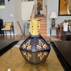 May include: A dark blue, diamond-shaped decorative oil lamp with gold filigree accents and a gold top. The lamp features ornate gold floral designs and is displayed on a gold surface.