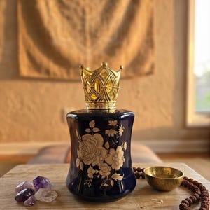 May include: A navy blue ceramic essential oil diffuser with a gold crown-shaped top. The diffuser features a floral design in gold. Amethyst crystals, a small brass bowl, and a beaded necklace are on a wooden surface.