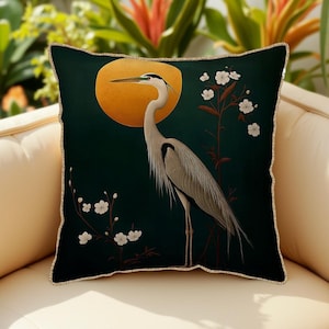 May include: A green throw pillow with a white heron standing in front of a yellow moon and white flowers. The pillow has a gold trim.