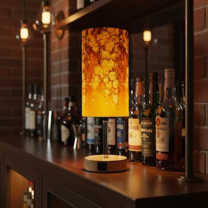 May include: A decorative table lamp with a cylindrical shade featuring a floral design in shades of yellow and brown. The lamp has a wooden base and a metal stem, sitting on a dark wooden surface. The background includes bottles and bar decor.