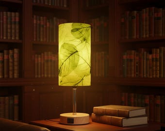 May include: A decorative table lamp with a cylindrical shade featuring a leaf pattern in shades of green and yellow. The lamp has a wooden base and a clear, slender stem. Two books are on the table. The background is a library with bookshelves.