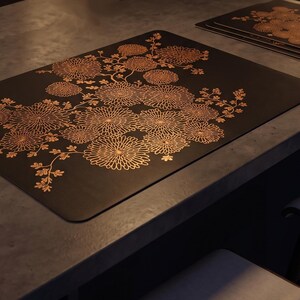 May include: A rectangular black placemat featuring a detailed copper-colored floral pattern of blooming chrysanthemums. The design covers the center and extends toward the edges of the mat, showcasing intricate petal lines and stems.