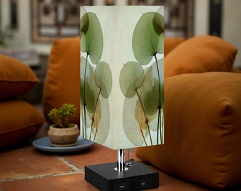 May include: A modern table lamp with a rectangular black base and a cream-colored shade. The shade features a repeating pattern of green and tan botanical illustrations. The lamp has a chrome stem and two USB ports.
