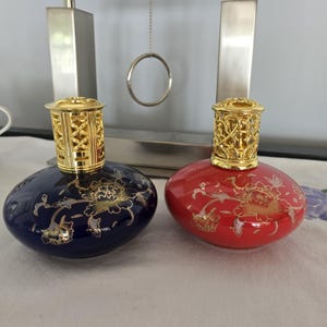 May include: Two round fragrance lamps, one dark blue and one red, each featuring a gold floral pattern and a decorative gold-tone metal lattice cap. The lamps have a smooth, glossy finish and a wide, flattened base design for stability.