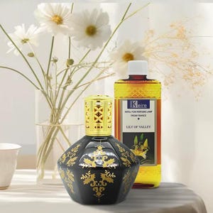 May include: A black and gold decorative fragrance lamp with a gold filigree top. A bottle of "Lily of the Valley" fragrance refill is behind the lamp. White flowers in a glass vase are in the background.