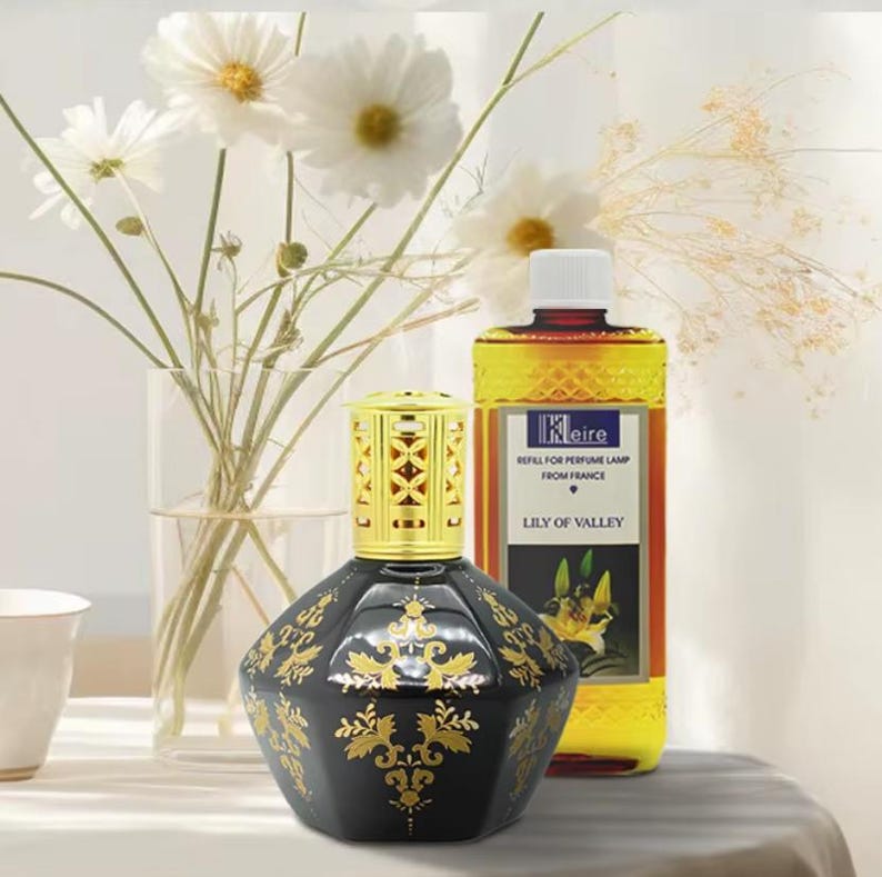 May include: A black and gold decorative fragrance lamp with a gold filigree top. A bottle of "Lily of the Valley" fragrance refill is behind the lamp. White flowers in a glass vase are in the background.