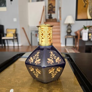 May include: A dark blue, diamond-shaped decorative bottle with gold floral designs. The bottle has a gold-colored, geometric top. The bottle is sitting on a gold-colored surface.