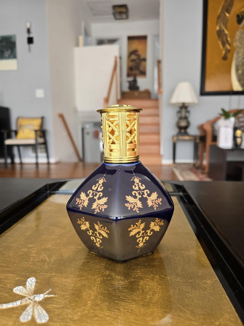 May include: A dark blue, diamond-shaped decorative bottle with gold floral designs. The bottle has a gold-colored, geometric top. The bottle is sitting on a gold-colored surface.