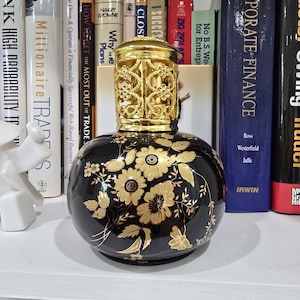 May include: A black, round decorative oil lamp with gold floral designs. The lamp has a gold filigree top and a glossy finish. The background includes a shelf with books.
