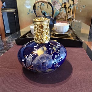 May include: A round, deep blue ceramic fragrance lamp featuring a gold-toned, openwork metal crown top. The body of the lamp is decorated with a painted floral pattern in shades of blue, gold, and white, depicting large blossoms and leaves.