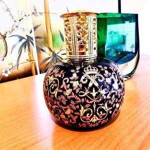 Pode incluir: Lâmpada de fragrância decorativa preta e dourada com uma parte superior de filigrana dourada. A lâmpada tem um design floral com detalhes em rosa e dourado. Um vaso de vidro verde está no fundo.