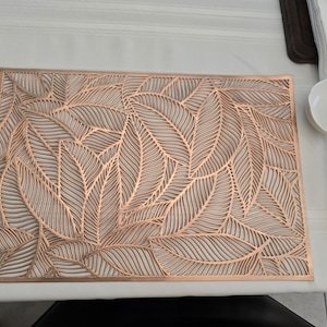 May include: A copper-colored placemat with a leaf design. The placemat is rectangular and features a cut-out pattern of overlapping leaves. A white bowl, chopsticks, and napkins are on the right side of the placemat.
