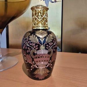 May include: A decorative black and gold fragrance lamp with a gold filigree top. The lamp features a floral design in gold and pink. The lamp is on a wooden surface.
