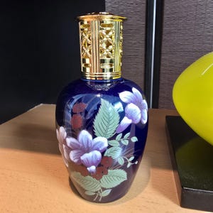 May include: A deep blue glass fragrance lamp with a glossy finish, featuring painted purple flowers, green leaves, and small red blossoms. The top of the vessel is fitted with a decorative, perforated gold-colored metal cap with a cross-hatch pattern.