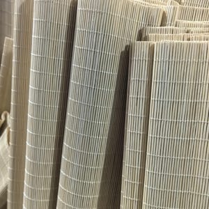 May include: A collection of light beige bamboo mats arranged in vertical layers. Each mat is constructed from thin, uniform bamboo slats held together by fine, light-colored thread, creating a textured, linear surface with a consistent pattern.