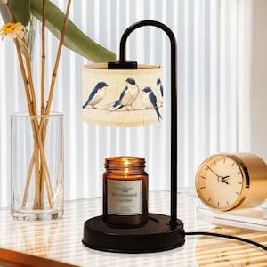 May include: A black metal candle warmer with a cylindrical lampshade featuring blue bird illustrations. A lit candle in a brown jar sits below the shade. A gold-colored clock is on the right.