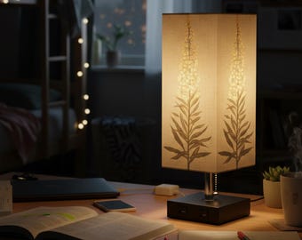 May include: A rectangular table lamp with a beige shade featuring a floral design. The lamp sits on a dark base with USB ports. The desk also holds a laptop, open book, smartphone, and writing supplies. The room has a bunk bed and string lights.