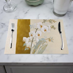 May include: A rectangular placemat with a floral orchid design. The placemat features a cream-colored background with a vertical gold stripe on the left side. A fork and knife are placed on the placemat.