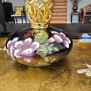 May include: A decorative black glass bottle with a gold crown-shaped lid. The bottle features floral designs in purple, red, and green. The bottle is displayed on a gold surface.