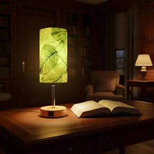 May include: A decorative table lamp with a cylindrical shade featuring a green leaf pattern. The lamp has a clear stem and a round wooden base. An open book rests on the dark wooden table, illuminated by the lamp's warm glow.