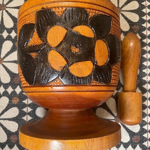 Könnte beinhalten: Ein Mörser- und Stößel-Set aus Holz mit einem geschnitzten Blumendesign. Der Mörser ist braun mit einem schwarzen Blumendesign und einem runden Boden. Der Stößel ist braun und hat eine glatte Oberfläche.